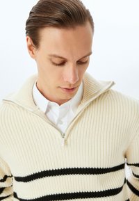 Crèmekleurige rits trui met geribbelde textuur en zwarte horizontale strepen. Heeft een staande kraag en een gelaagd uiterlijk over een wit shirt.