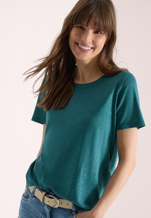 Femme souriante aux longs cheveux bruns portant un t-shirt à manches courtes couleur sarcelle et une ceinture beige clair avec un jean bleu sur un fond clair.