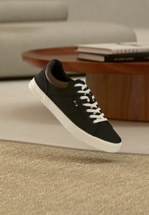 Schwarzer Sneaker mit niedrigem Schaft, weißen Schnürsenkeln und weißer Sohle, schwebt über beigem Teppich, im Hintergrund ein Holztisch und übereinander gestapelte Bücher.