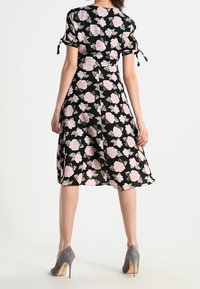 Frau in einem schwarzen Midi-Kleid mit pinkem Blumenmuster und Bindedetails an den kurzen Ärmeln, kombiniert mit grauen High Heels, steht mit dem Rücken zur Kamera.