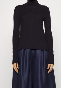 Pull à col roulé noir côtelé à manches longues, en tissu tricoté. L'ourlet est droit et légèrement plus long à l'arrière.