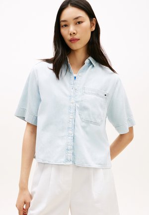 Νεαρή γυναίκα με μακριά σκουρόχρωμα μαλλιά, ντυμένη με γαλάζιο πουκάμισο με κοντά μανίκια και λευκό παντελόνι, στέκεται μπροστά σε απλό φόντο.