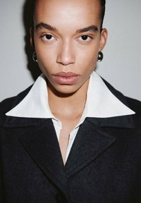 Abrigo negro con un gran cuello blanco, que presenta una tela texturizada. Primer plano de una persona con maquillaje minimalista y pendientes oscuros.