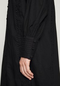 Vestido preto com um placket frontal com babados, punhos estruturados com botões e um tecido suave e texturizado. Vista lateral destacando o detalhe das mangas.