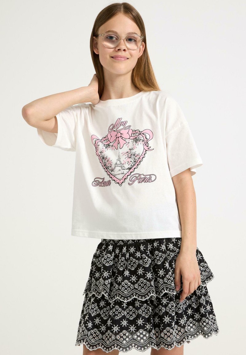 Vitt kortärmat t-shirt med en rosa grafisk hjärtformad design föreställande ett Eiffeltorn. Svart lagrad kjol med vita blommönster och spetsdetaljer.