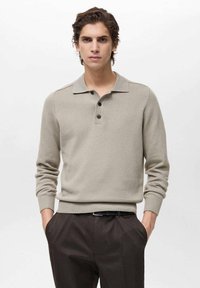 Beige knitted polo shirt with a collared neckline, three black buttons, and ribbed cuffs. Paired with dark trousers for a classic look.