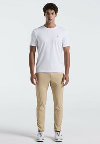 Witte korte mouwen t-shirt met klein logo op de borst, gecombineerd met beige slim-fit broek en witte sneakers op een effen achtergrond.