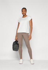Haut de sport blanc à manches courtes, leggings marron et baskets blanches. Un sac de sport noir est tenu dans une main. Textures lisses et design ajusté.