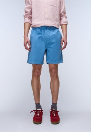 Mężczyzna w różowej koszuli w paski z długim rękawem, niebieskich szortach, szarych skarpetach i czerwonych trampkach stoi na jednolitym tle.