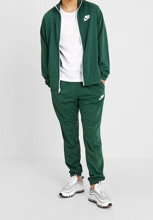 Person trägt dunkelgrünen Nike-Trainingsanzug mit weißem T-Shirt und weißen Nike-Sneakern vor neutralem hellen Hintergrund.