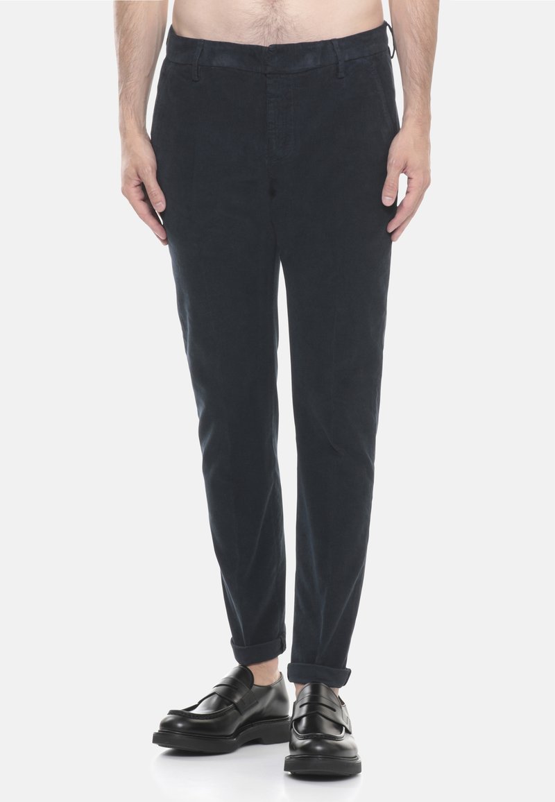 Uomo che indossa pantaloni in velluto a coste neri arrotolati alle caviglie e mocassini in pelle nera, in piedi contro uno sfondo semplice.