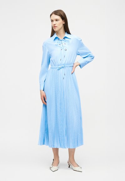 Γυναίκα που στέκεται με το χέρι στη μέση, φοράει ένα μακρυμάνικο, γαλάζιο ριγέ μίντι φόρεμα με ζώνη στη μέση και λευκά παπούτσια με χαμηλό τακούνι.