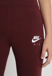 Vinröda leggins i stretchmaterial med en vit Nike Air-logga på vänster lår och en slät, stilren textur.