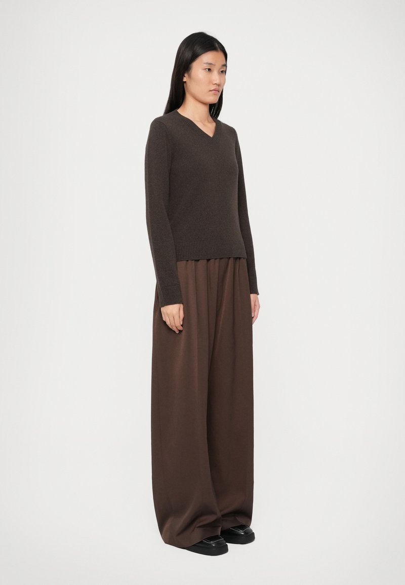 Woman wearing a dark brown V-neck sweater and wide brown pants standing against a plain light background, facing slightly right.
