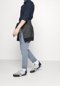 Avslappnad outfit med en marinblå skjorta, ljusblå byxor och grå sneakers med marinblå och svarta detaljer, kompletterad med en mörk stickad tröja knuten runt midjan.