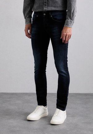 Man draagt donkerblauwe slim-fit jeans, witte sneakers en een grijs overhemd, staand op een grijze vloer.