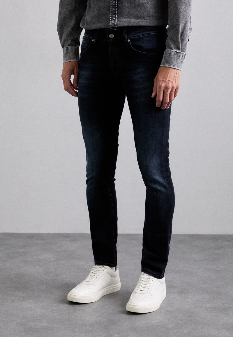 Homme portant un jean slim bleu foncé, des baskets blanches et une chemise grise boutonnée debout sur un sol gris.