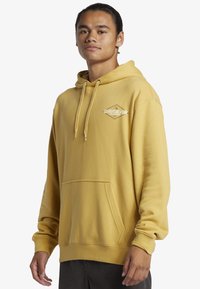 Yellow hooded sweatshirt made of soft fabric, featuring a front pocket, drawstring hood, and a logo in a diamond shape on the chest.