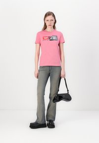 Rosa bomull t-skjorte med grafisk logo, sammen med lysegrå snekkerjeans og svarte platåsko. En svart håndvogn bæres av modellen.