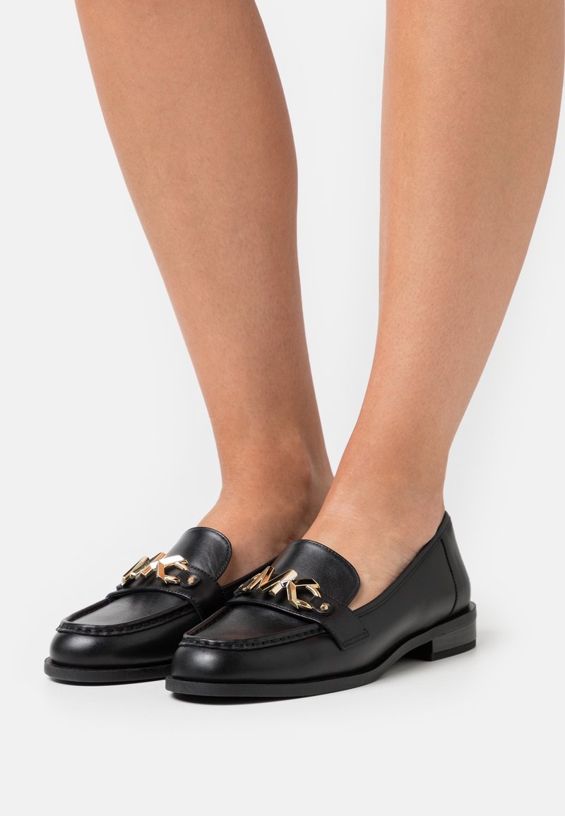 Mocassins en cuir noir avec bout arrondi, ornés d'une chaîne dorée et d'un talon plat. Texture lisse et design simple avec coutures discrètes.