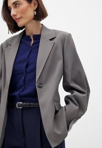 Pilkas švarkas su struktūrizuotu dizainu, turintis platų apykaklės kraštą ir vieną sagą. Dėvimas ant tamsiai mėlynos marškininės su saga, derinamas su juodu diržu.