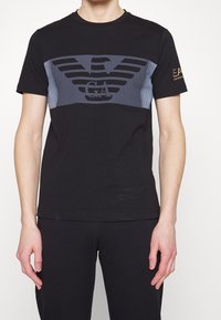 Svart bomulls-t-shirt med en grå grafisk design med vingar och logotyp, korta ärmar och rund hals. Accenterad med varumärkestext på ärmen.