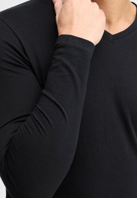 Juoda ilgų rankovių V kaklo marškinėliai, pagaminti iš lygaus audinio; prigludusi konstrukcija pabrėžia rankas ir pečius, su švariu, minimalistiniu įvaizdžiu.