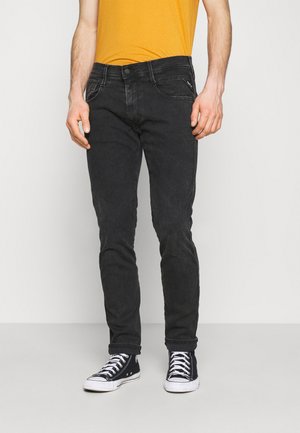 Homme portant un jean noir ajusté, une chemise jaune et des baskets montantes noires, debout devant un fond clair uni.