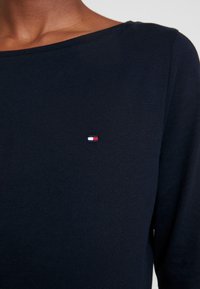 Navy pitkähihaine paita pehmeästä kankaasta. Rinnassa on pieni brodeerattu logo punaisena, valkoisena ja sinisenä. Tasainen pääntie.