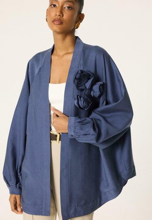 Femme portant une veste bleue ample avec des détails de fleurs en tissu sur la poitrine, par-dessus un haut blanc et un pantalon beige, debout devant un fond uni.