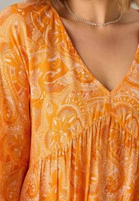 Mujer vistiendo un vestido naranja con patrón paisley y escote en V, con collar de cadena plateada, mostrando la parte superior del pecho y los hombros.