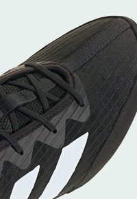 Nærfoto af sort atletik sko med tekstureret stof, sorte snørebånd og to markante hvide diagonalstriber på siden.