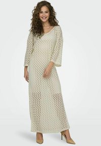 Vestido de crochet largo de color claro con escote en V, mangas tres cuartos y ajuste holgado. Lleva puestos unos tacones altos beige.