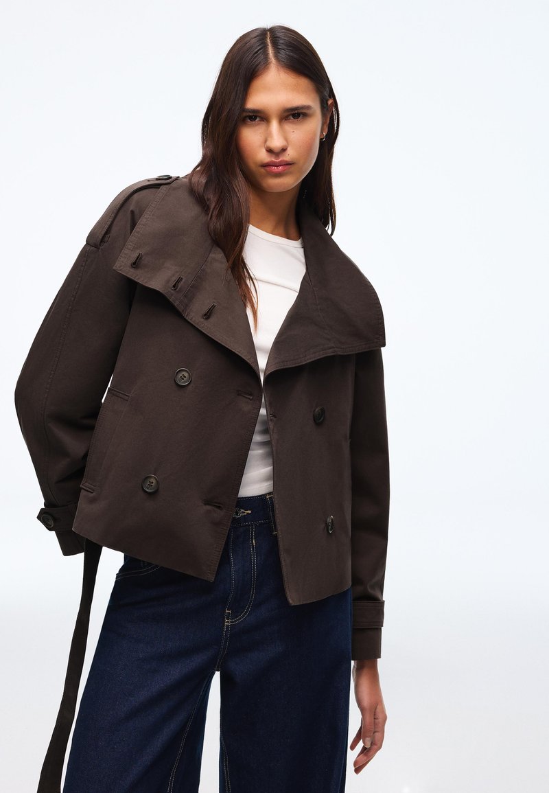 Giovane donna che indossa un trench corto marrone scuro sopra una camicia bianca e jeans a vita alta blu scuro, in piedi contro uno sfondo semplice.