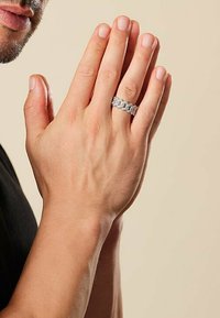 Silverring med flätad design, bärs på en hand med fingrarna samlade. Hudtonen är naturlig med lätt textur. Neutral bakgrund.