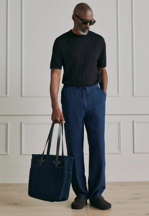 Mand iført sort kortærmet sweater, marineblå bukser med snor i taljen, sorte sko og solbriller, der holder en stor marineblå mulepose med sorte stropper.