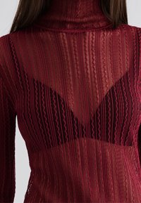 Haut à col roulé en rouge translucide avec un motif texturé à rayures verticales et une superposition de soutiens-gorge triangulaire noire contrastante.