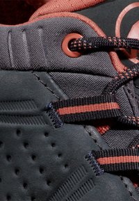 Scarpa sportiva nera con tessuto testurizzato e perforazioni. Presenta lacci scuri fissati da un occhiello rotondo; accenti rosa lungo la parte superiore.