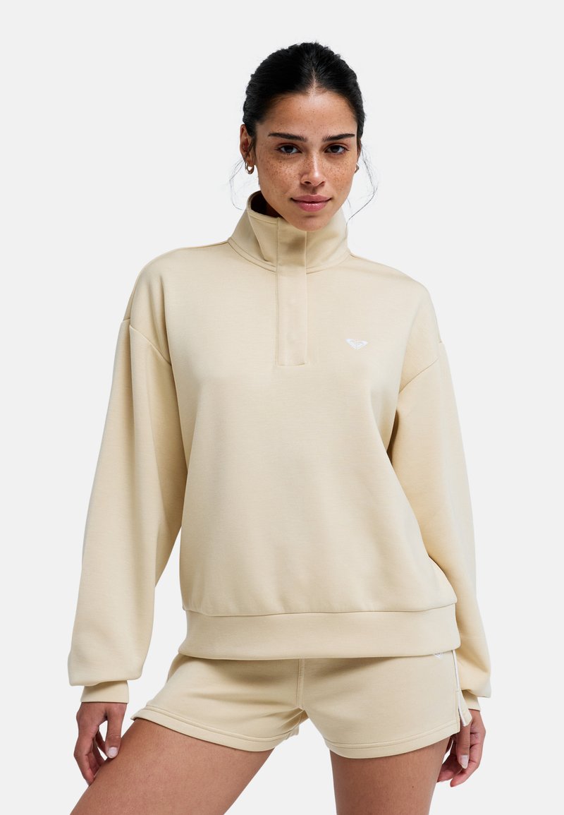 Sudadera extragrande beige con cuello alto y media cremallera, con un pequeño logo blanco. Combinada con pantalones cortos beige a juego. Textura suave.
