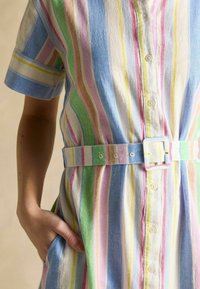 Gestreiftes Kleid in Pastellfarben, mit Knöpfen, einem taillierten Taillengürtel, kurzen Ärmeln und einer lockeren Passform. Hergestellt aus leichtem Stoff.