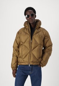 Giacca puffer marrone con design a rombi, polsini elasticizzati e cappuccio con cordino. Abbinata a jeans scuri e occhiali da sole.