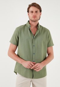 Cămașă cu mânecă scurtă, cu guler, dintr-un material verde deschis, având nasturi albaștri, o suprafață texturată și o margine dreaptă.