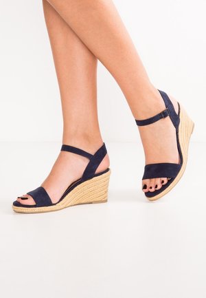 Ben iført marineblå ruskind kilehæls-sandaler med ankelstropper og teksturerede beige såler, tæer malet sorte, på hvid baggrund.