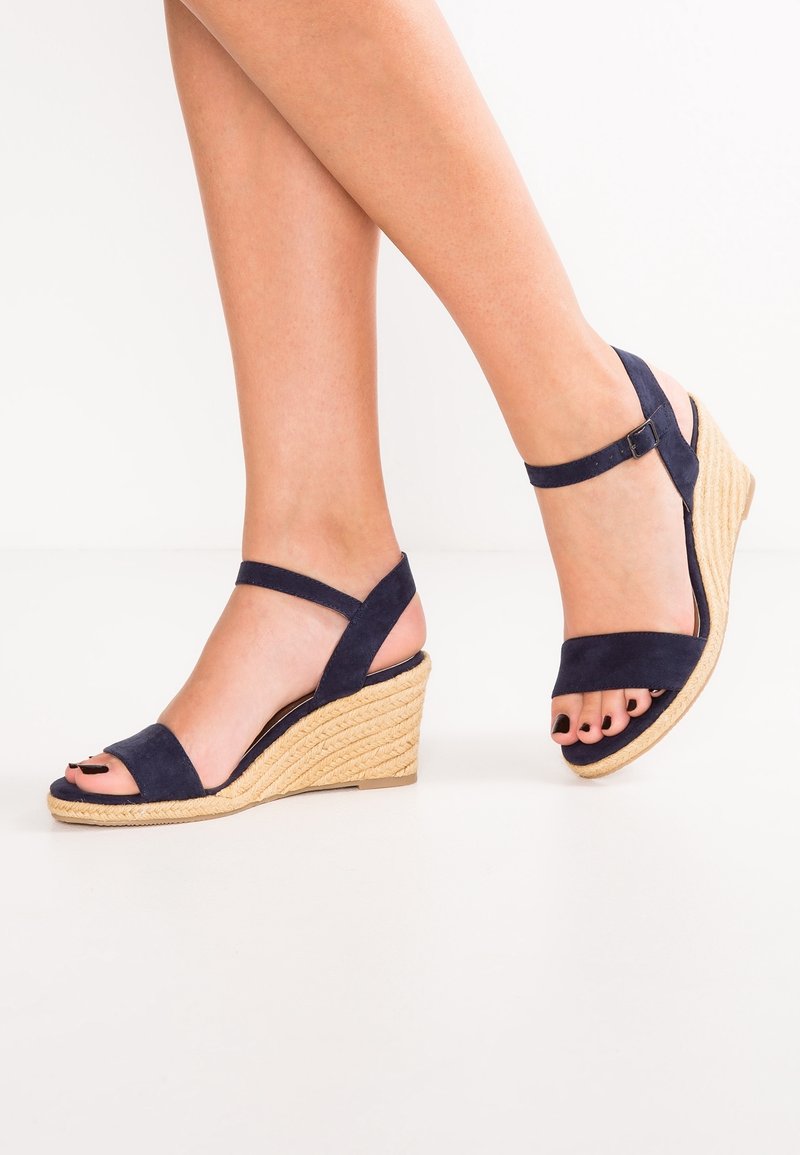 Jambes portant des sandales compensées en daim bleu marine avec lanières à la cheville et semelles texturées beiges, orteils peints en noir, sur fond blanc.