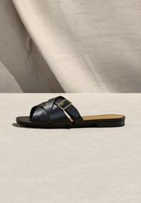 Sandale plate noire à enfiler avec des lanières en cuir tressé et une boucle dorée sur une surface neutre et un fond en tissu beige.