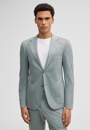 Mann trägt hellgraues Sakko und passende Hose über weißem Rundhalsausschnitt vor schlichtem Hintergrund.