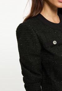 Svart texturerad jacka med ett glänsande tråd-mönster, rund halsringning och en metallknappdetalj på vänster sida.