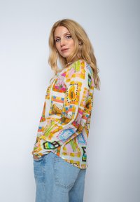 Buntes Langarmshirt mit einem lebhaften, gemusterten Design, das Elemente in Pink, Grün, Orange und Gelb auf einem hellen Hintergrund zeigt.