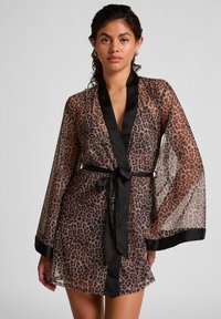 Vrouw in een doorschijnende shirt met luipaardprint en zwarte afwerking, met een zwarte tailleband, staande tegen een effen lichte achtergrond.