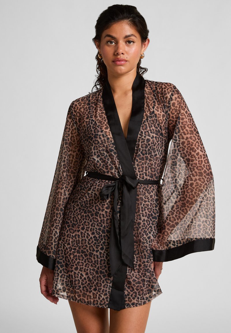 Vrouw in een doorschijnende shirt met luipaardprint en zwarte afwerking, met een zwarte tailleband, staande tegen een effen lichte achtergrond.